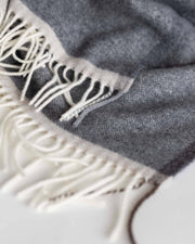 Close-up of a gray herringbone woven textile with off-white fringe along one edge, photographed on a light neutral background.