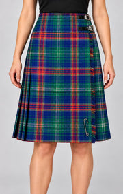 A woman wearing a pleated kilt skirt in a blue, green, red, and yellow tartan plaid pattern, featuring a black belt and a silver safety pin detail, shown against a solid gray background.