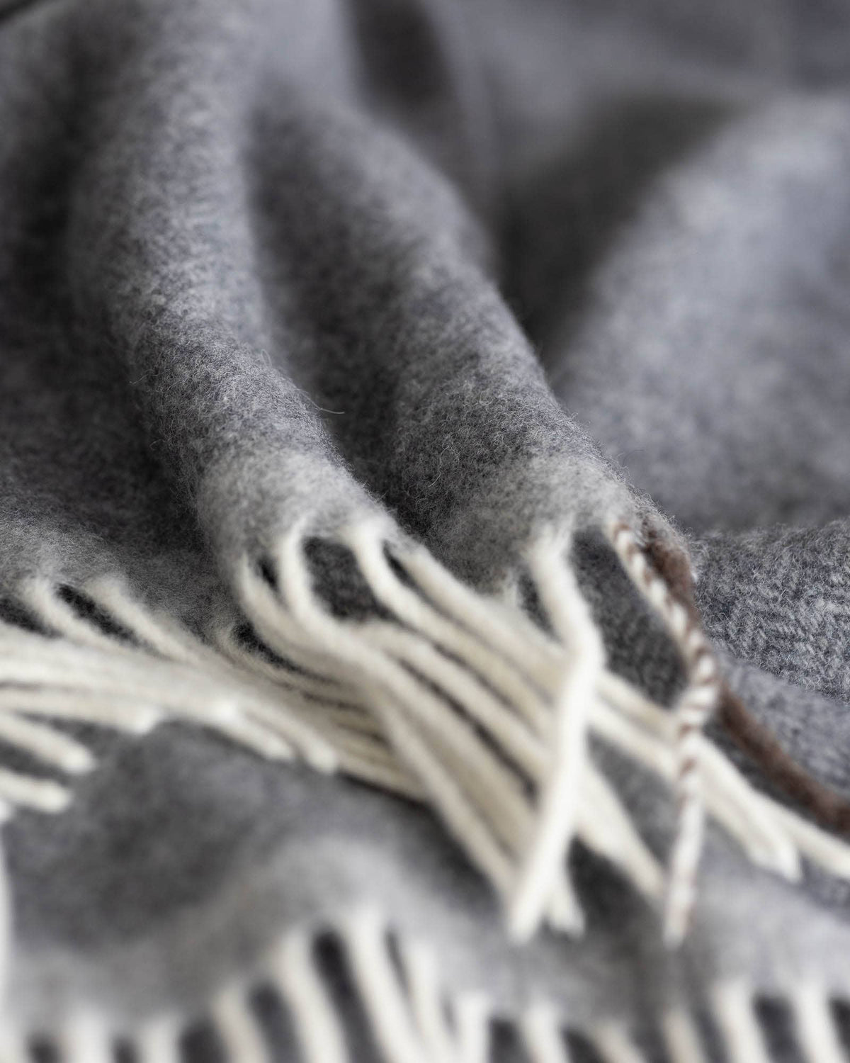 Close-up of a soft gray woven throw blanket with cream fringe and a small brown embroidered accent near the edge.